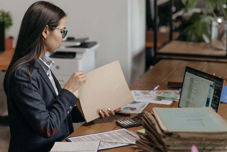 Una mujer con lentes y saco oscuro revisa una carpeta en su escritorio, junto a una pila de expedientes, una calculadora y una laptop con una hoja de cálculo