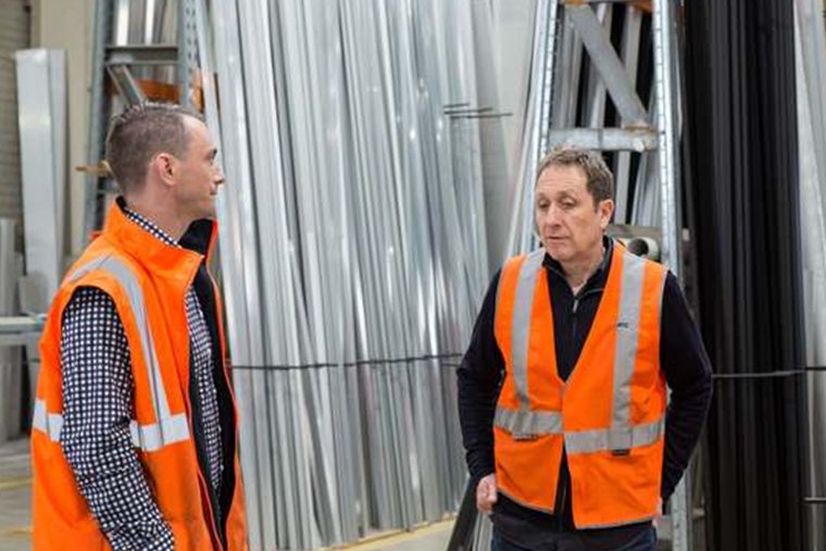 Tres hombres con chalecos reflectivos conversan de pie en el almacén de una empresa industrial, entre estanterías con perfiles de aluminio