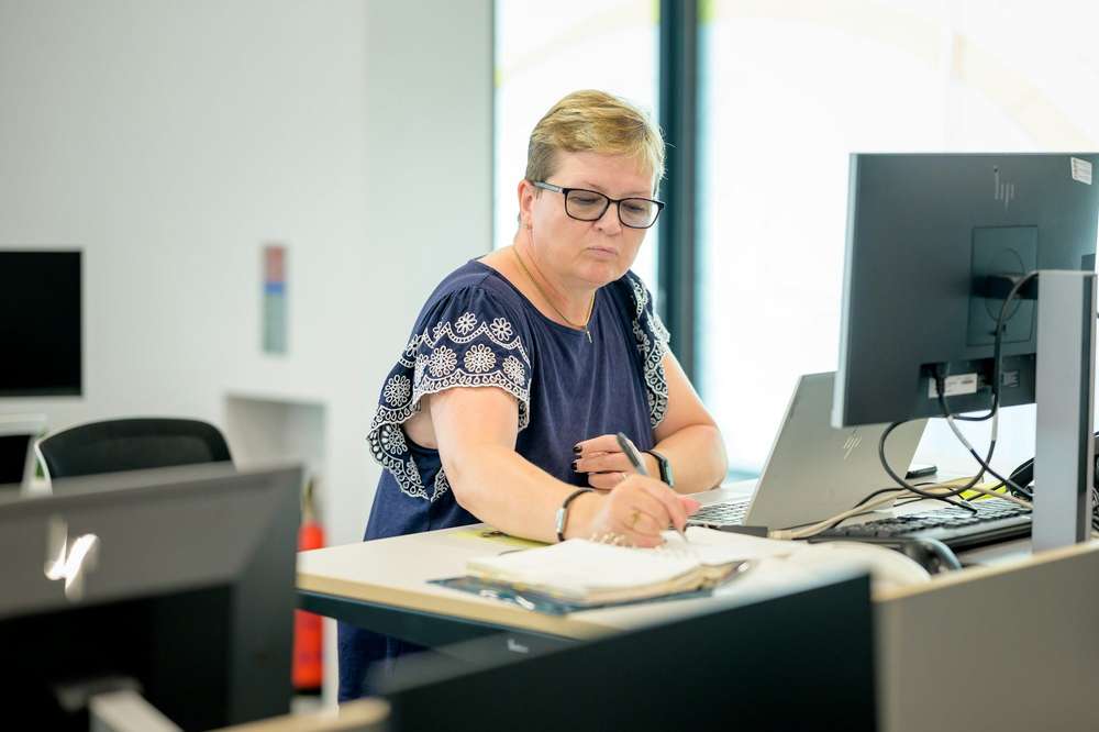 Una mujer con lentes, de pie en su puesto de trabajo, anota en su agenda junto a una laptop y un monitor, en una oficina luminosa