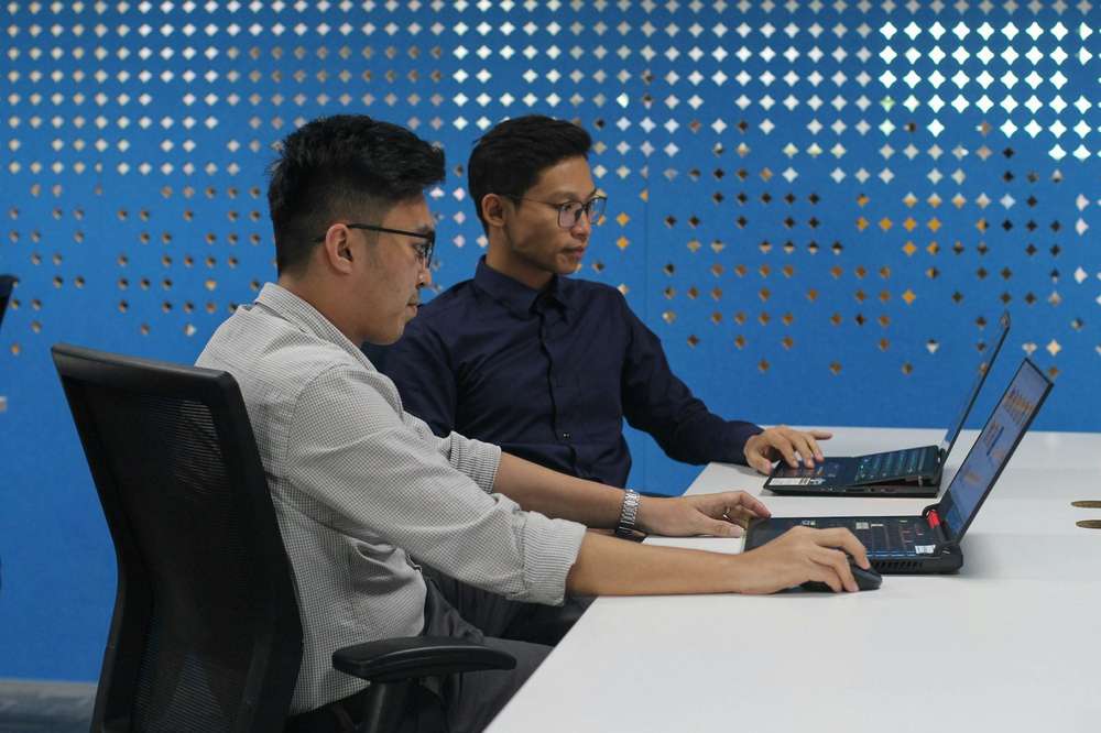 Dos hombres con lentes trabajan lado a lado en sus laptops, en una mesa blanca frente a una pared azul perforada
