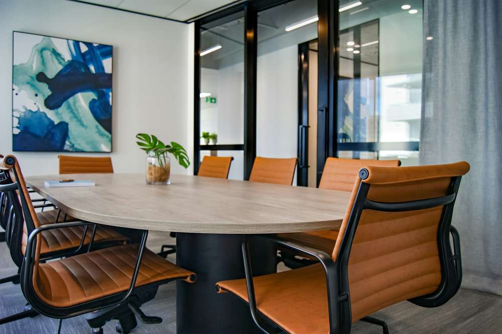 Una sala de reuniones vacía, con una mesa ovalada de madera, sillas color coñac, un cuadro azul abstracto y paredes de vidrio