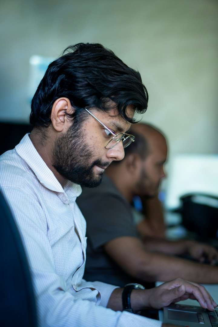 De perfil, un hombre de barba y lentes escribe concentrado en su laptop; a su lado, desenfocado, un compañero trabaja en la suya