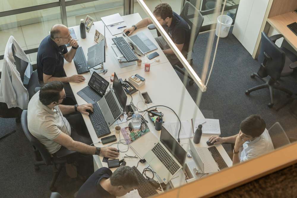 Vista desde arriba, a través de un vidrio: cinco personas trabajan en laptops y teclados alrededor de una mesa larga llena de cuadernos, cables y papeles