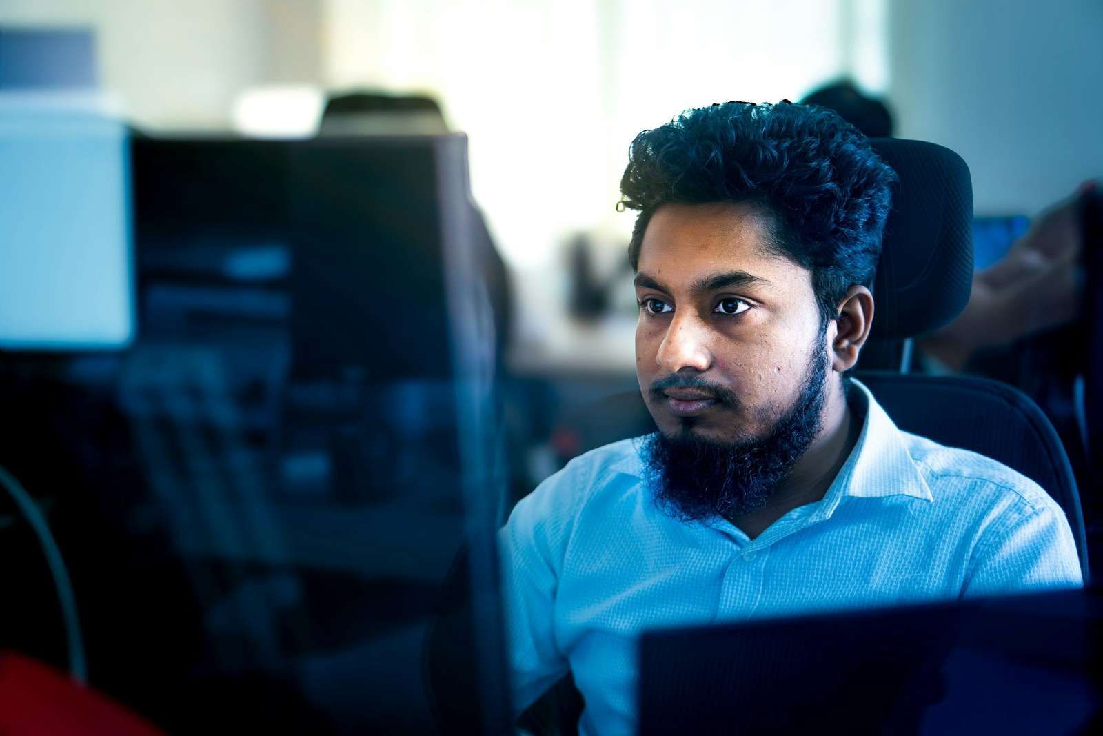 Un hombre de barba y camisa celeste trabaja concentrado frente a su monitor, en una oficina con más puestos de trabajo desenfocados detrás
