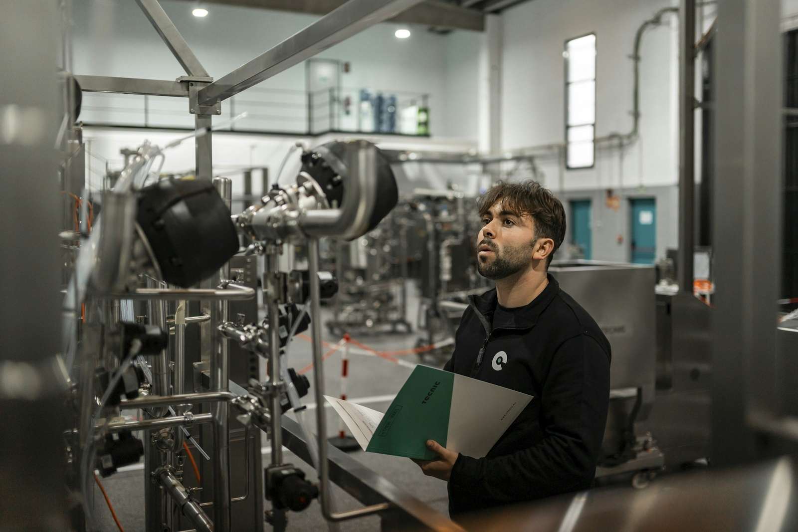Un técnico con una carpeta en la mano revisa la maquinaria de acero inoxidable de una planta industrial moderna y limpia