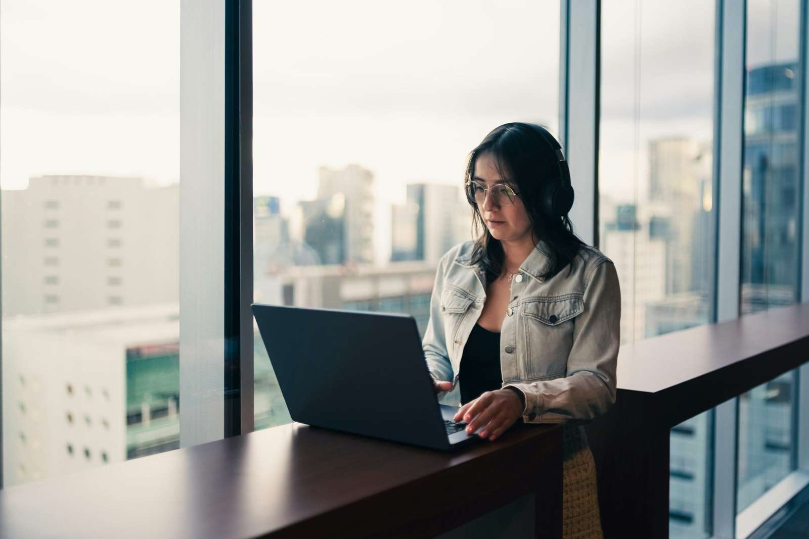 Una mujer con lentes y audífonos trabaja de pie en su laptop, junto a un ventanal de piso a techo con edificios de la ciudad detrás